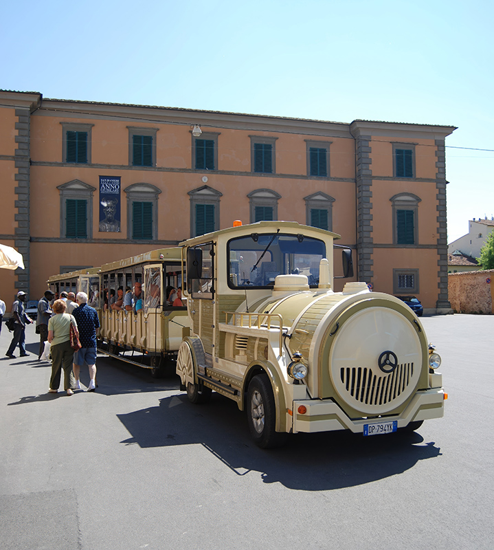 3 - A beige and cream-colored tourist trackless train (road train) parked on a paved square in front of an historic orange-facade building in Italy